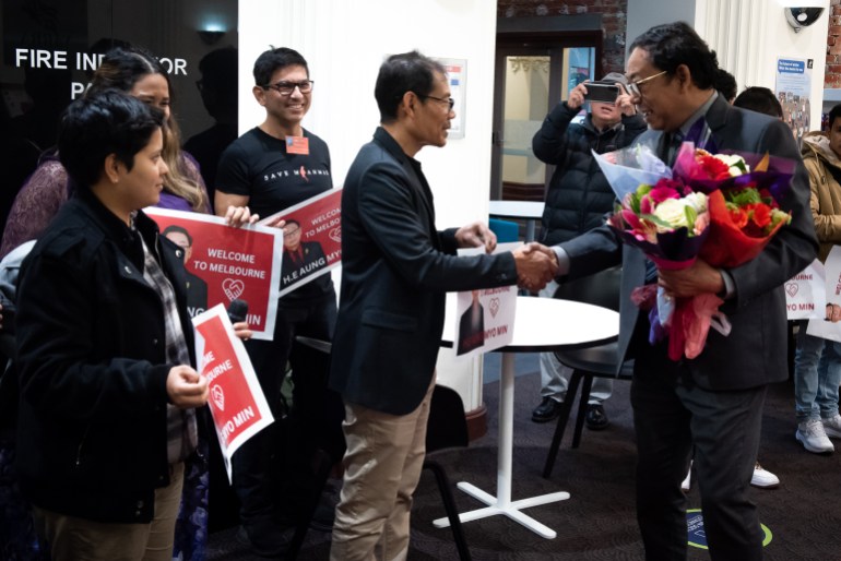NUG Human Rights Minister Aung Myo Min shaking hands with community members in Melbourne. He is holding a bouquet of flowers. Some people are holding placards reading Welcome to Melbourne. Everyone looks happy.