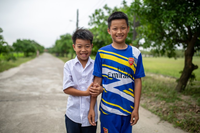 Two boys who found an unexploded weapon when they were playing near a muddy ditch. They are standing on road. The shorter boy is hanging onto the taller boy's arm. They are both smiling.