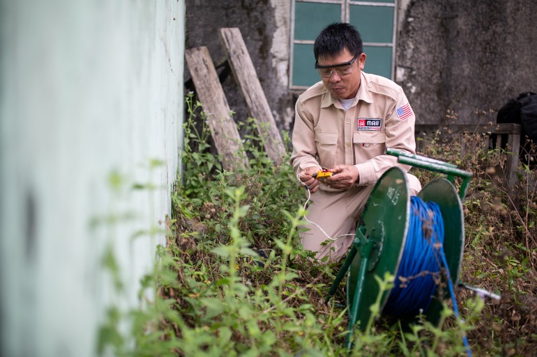 An expert prepares to destroy cluster munitions and a grenade found in a Vietnamese village. He is squatting down near a wall and wearing protective glasses.