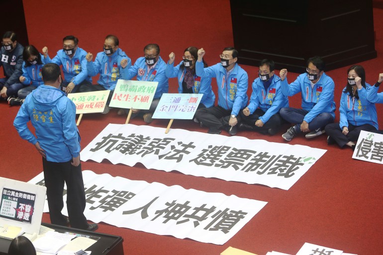 KMYT legislators dressed in sky blue bomber jackets sitting on the floor of the legislature in protest against the anti-infiltration bill. They have banners in Chinese characters reading 'Protest against a bad law", "Sanction by Votes. "Neck Bomb" "Be hated by both man and God''. The banners are laid out on the floor. Three are holding a banner in their hands.