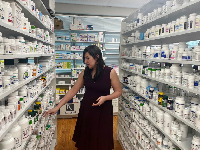 A woman walks through the aisles of a pharmacy, with shelves full of medication.