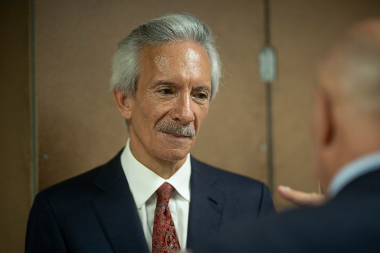 A close-up of an older man with a mustache in a dark suit and red tie.
