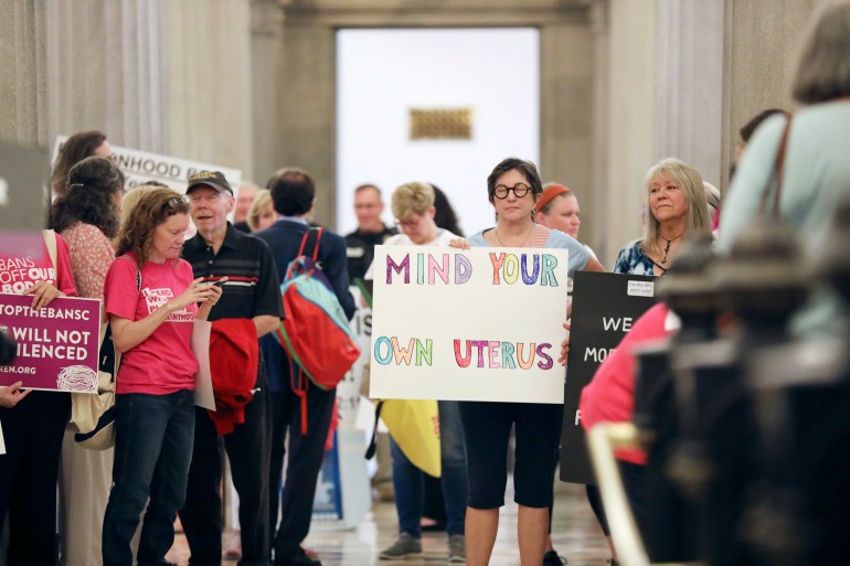 A photo of a group of people protesting and holding signs with the person in the middle holding a sign that says "mind your own uterus".