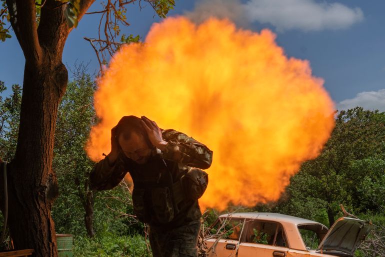A Ukrainian soldier covers his ears and ducks after firing a mortar. The gun is behind him and there is a huge orange cloud of smoke