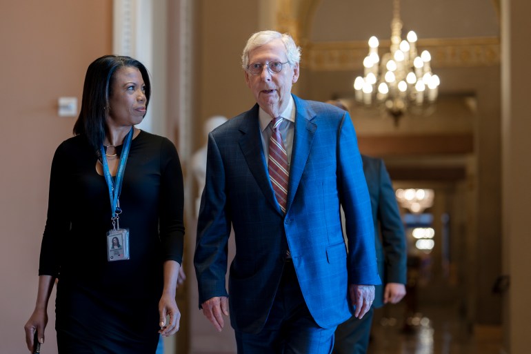 Mitch McConnell walks through the halls of Congress in a blue suit, accompanied by a colleague