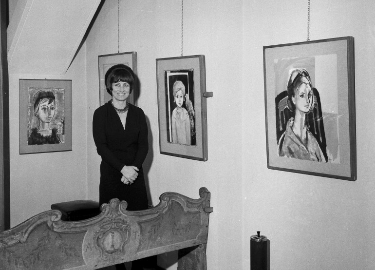 Francoise Gilot posing with her work at her exhibition in Milan in 1965. The photo is black and white. She is smiling and looks very happy. The paintings are portraits.