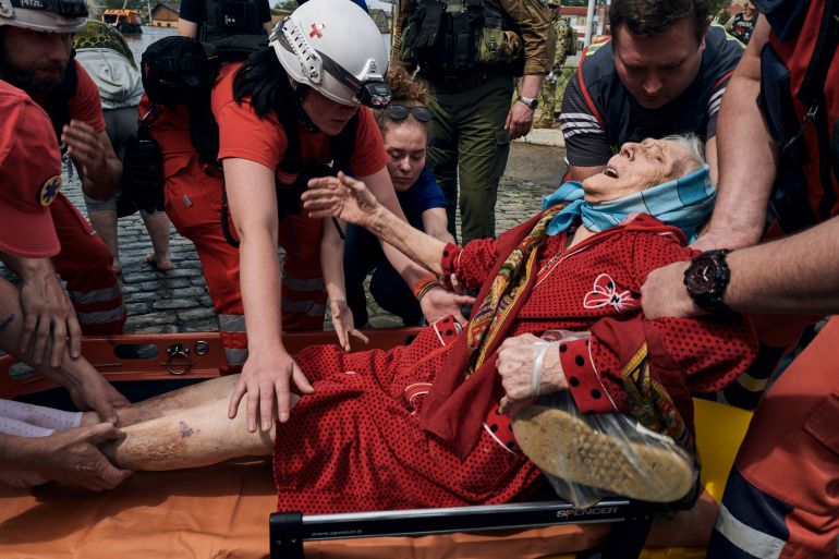 Am elderly woman being helped onto a stretcher by rescuers during Kherson flood evacuations. She is in a red robe and has a blue scarf tied around her head and appears to be in pain