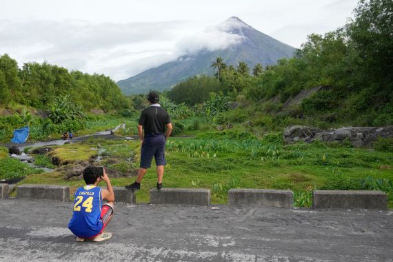 A boy takes a photo of his friend who poses in front of the Mayon volcano