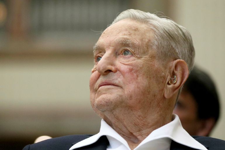 A portrait of George Soros. He is wearing a white open-necked shirt and black jacket. He is listening to someone speaking.