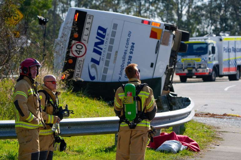 Three firefighters standing at the side of the road behind the overturned bus. The bus is lying on its side. There is a fire engine to the right
