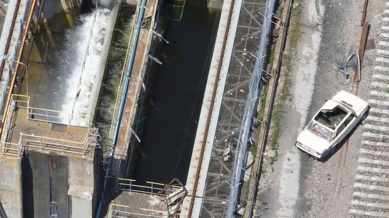A drone photo showing the top of the Nova Kakhovka dam with an old cream coloured car parked on top. The roof of the car is missing and there appear to be two large barrels inside.