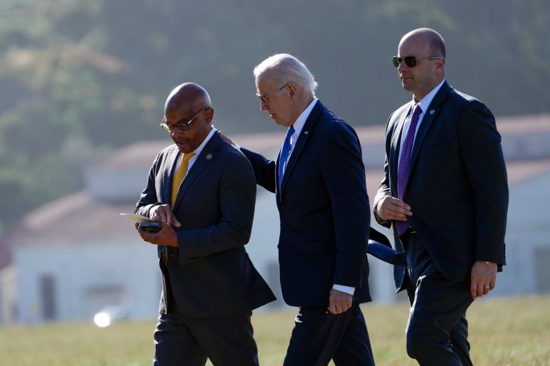 Joe Biden walking with an aide. He is patting the aide on the shoulder. Biden is wearing his trademark aviator-style sunglasses.