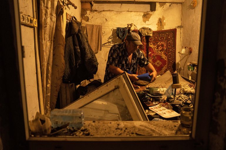 A man picking up important documents in his home. The flat has been hit by a Russian missile so the windows are blown out, there is debris over the floor and no power.
