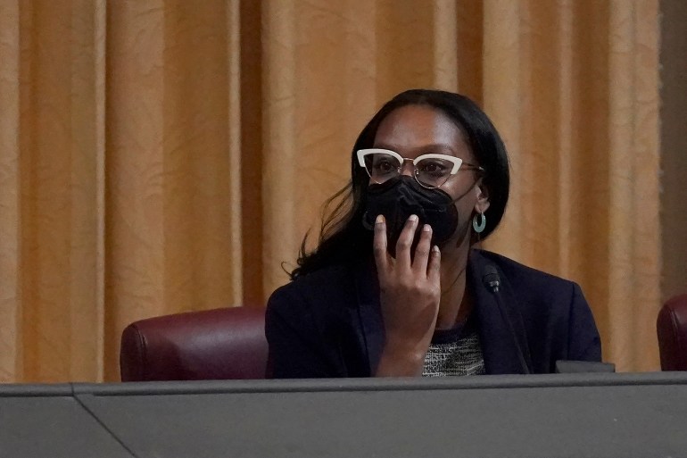 Seorang wanita, mengenakan masker dan kacamata, membungkuk di atas meja panel dan mendengarkan.