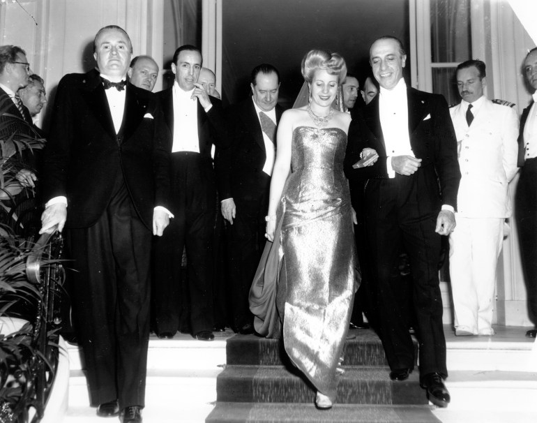 A woman in a shiny gown walks arm-in-arm with a man in a white tie as they travel down a set of steps