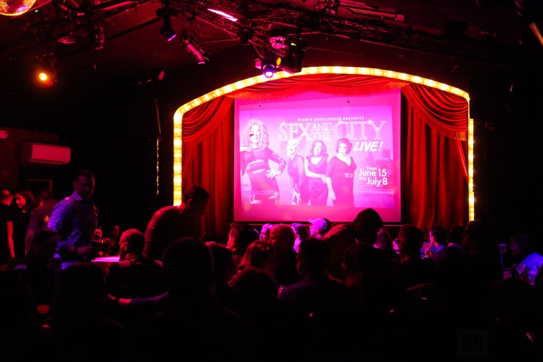 Inside the Oasis, for a cabaret style setup "Sex and the City Live," An advertisement for which is presented on a screen on a stage.