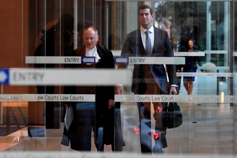 Ben Roberts-Smith keluar dari pintu kaca Pengadilan Federal Australia di Sydney pada Juni 2021 setelah mengajukan kasus pencemaran nama baik terhadap tiga surat kabar.  Dia mengenakan setelan gelap dan memakai jas.  Seorang pengacara berjubah ada di depannya