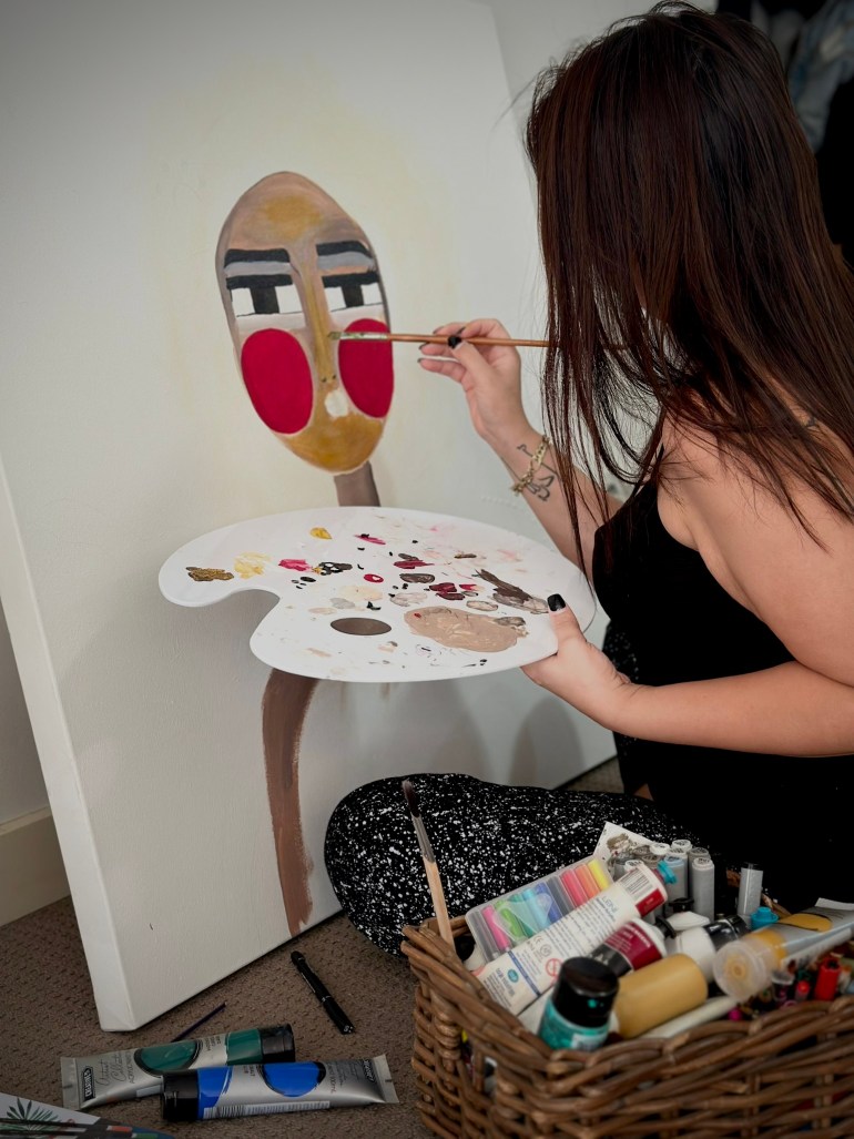 Maya arbeitet an einem ihrer Gemälde.  Es zeigt eine Figur mit leuchtend roten Wangen.  Sie hält eine Palette in ihrer linken Hand, während sie an der Leinwand arbeitet.