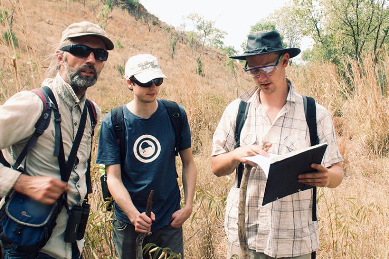 Jochen Brocks and Benjamin Nettersheim, exploring outcrops in northern Australia. They're dressed for hot weather and looking at a file. There is undergrowth and an escarpment behind them