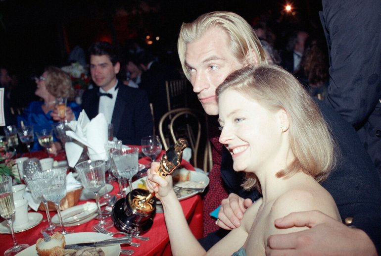 Jodie Foster, a grin on her face and her Oscar in hand, sits with Julian Sands during the Governor's Ball at the Shrine Auditorium on March 29, 1989. Sands has his arm around her.