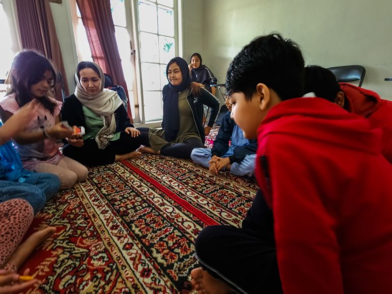 Afghan refugee children in Indonesia. They are seated on a carpet on the floor. One girl has a pack of Uno cards in her hands and they are about to play. They look excited.