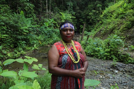 A portrait of Yustina Ogoney. SHe is standing in the forest wearing a red dress with a yellow necklace. She has a blue headband.