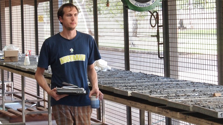 Geochemist Benjamin Nettersheim studying sample cores from the rocks. He's dressed casually and carrying some materials. The samples are laid out in trays on tables beside him.