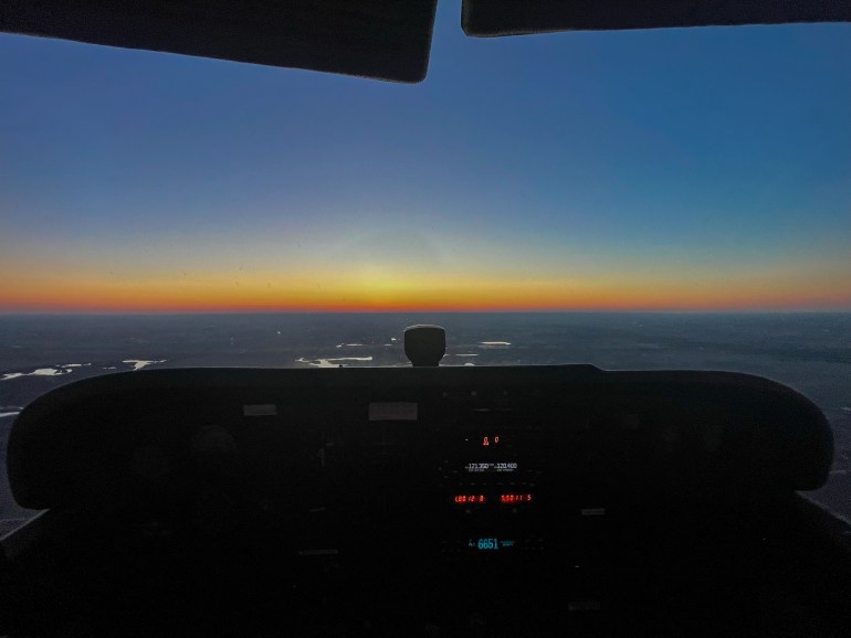 A photo of the pilot's view at sunrise of the controls and window in a small plane with the ocean below.