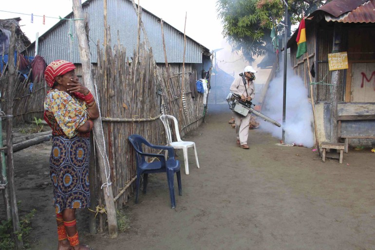Seorang wanita berdiri di dekat pagar, sementara petugas kesehatan bertopeng berjalan di sekitar rumah penduduk setempat, dengan alat yang mengeluarkan asap tebal.