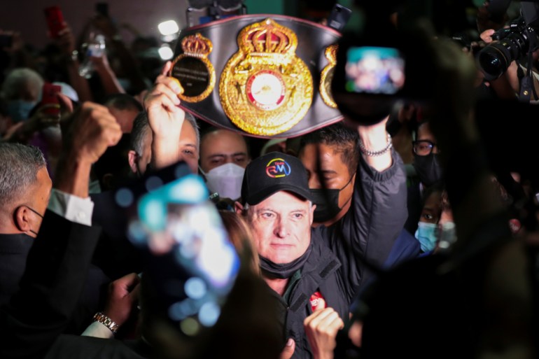 A man, amid a crowd, lifts a big black boxing belt with a red crest in the center