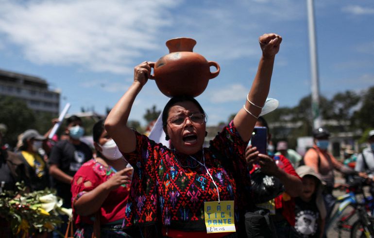 An Indigenous woman in an embroidered garment raises a fist as she balances a clay pot full of water on her head.