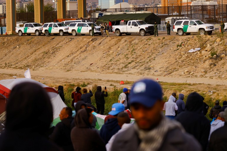 A row of pickup trucks can be seen parked along one bank of the Rio Grande river, as migrants and asylum seekers gather on the other side.