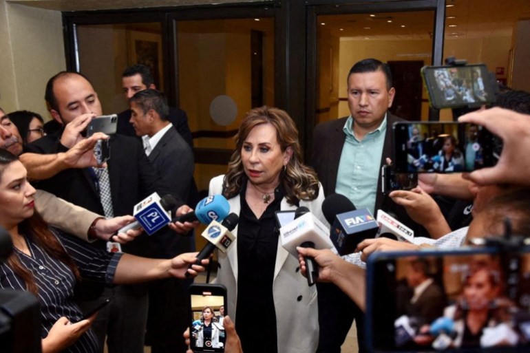 Seorang wanita berdiri dikelilingi oleh wartawan yang memegang mikrofon ke arahnya.