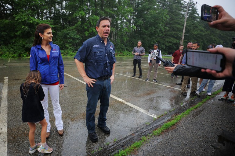 A man in a blue collared shirt stands next to his wife and small child on the concrete, as reporters hold microphones out to him. The concrete is wet from recent rain.