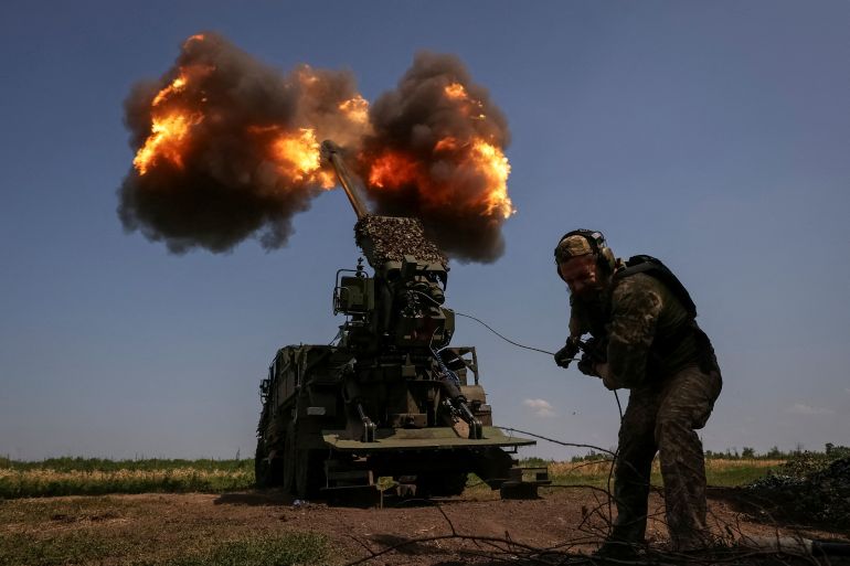 Ukraynalı bir asker, Bakhmut yakınlarındaki Rus hatlarına obüs ateşlerken eğiliyor.  Silah arkasında ve duman ve alev bulutları var