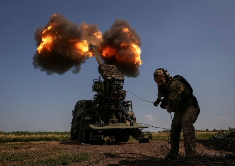 A Ukrainian soldier ducks as he fires a howitzer towards Russian lines near Bakhmut. The weapon is behind him and there are clouds of smoke and flames