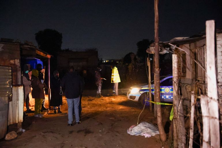 Police stand at a darkened scene at night, where improvised houses can be seen with panels of scrap metal and wood. A police vehicle is parked nearby.