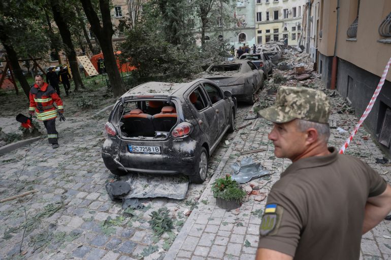 The damage after a Russian missile hit a residential building in Lviv. There is dust and rubble on the street. Cars have been destroyed and trees brought down. An official is standing to the right of the photo looking at the damage. He is wearing khaki coloured clothing and a hat, with a Ukrainian flag on the sleeve of his t-shirt.