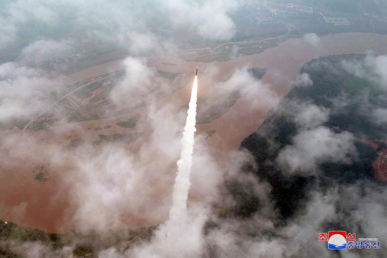 A wide aerial view of the missile as it climbs through the clouds. There is a wide belt of muddy brown river below.