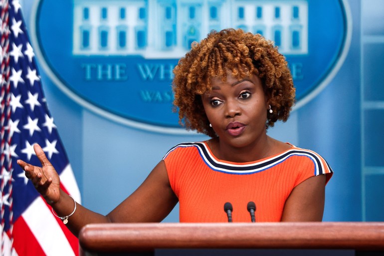 A woman in an orange top stands behind a podium outfitted with a microphone. Behind her is an American flag and the seal for the White House.