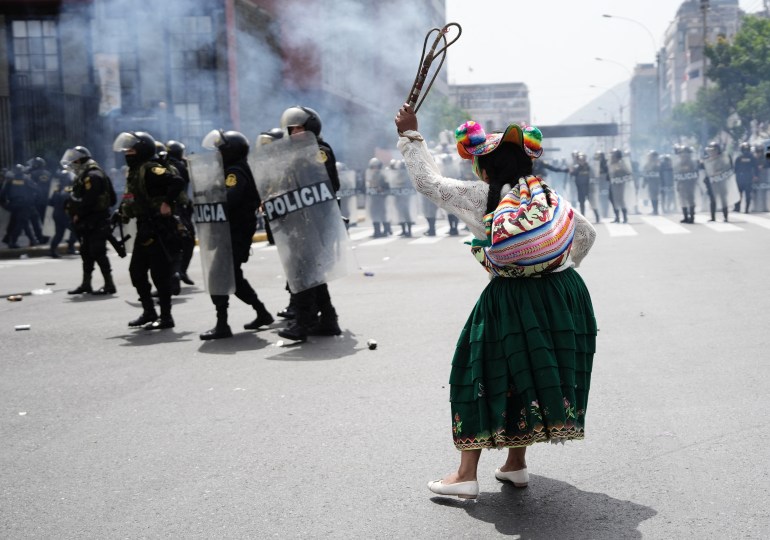 Security forces in riot gear — plastic shields, helmets, flak jackets — march through the streets as a demonstrator in traditional clothing waves her hand in the air.