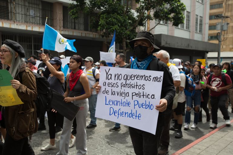 Enquanto as pessoas marcham, agitando bandeiras da Guatemala, um homem segura uma placa manuscrita.