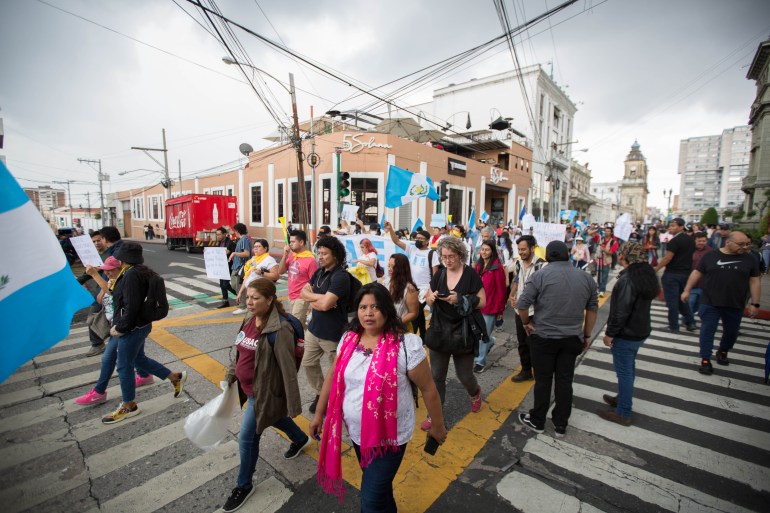 Группа людей, некоторые с написанными от руки табличками и гватемальскими флагами, маршируют через перекресток под серым грозовым небом.