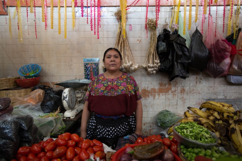 A woman in an embroidered top stands behind a table brimming with fruits and vegetables, including tomatoes and bananas. Behind her, garlic hangs from a wall.