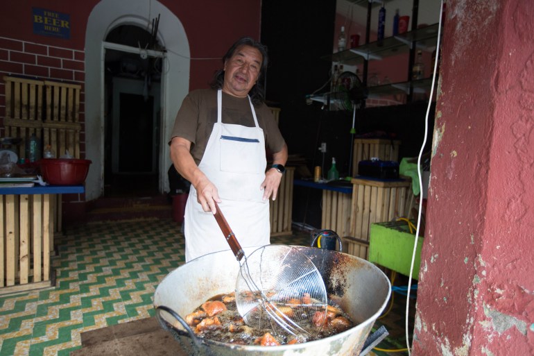 A man in a white apron stirs a large metal pot filled with pork rinds in the doorway of a shop.