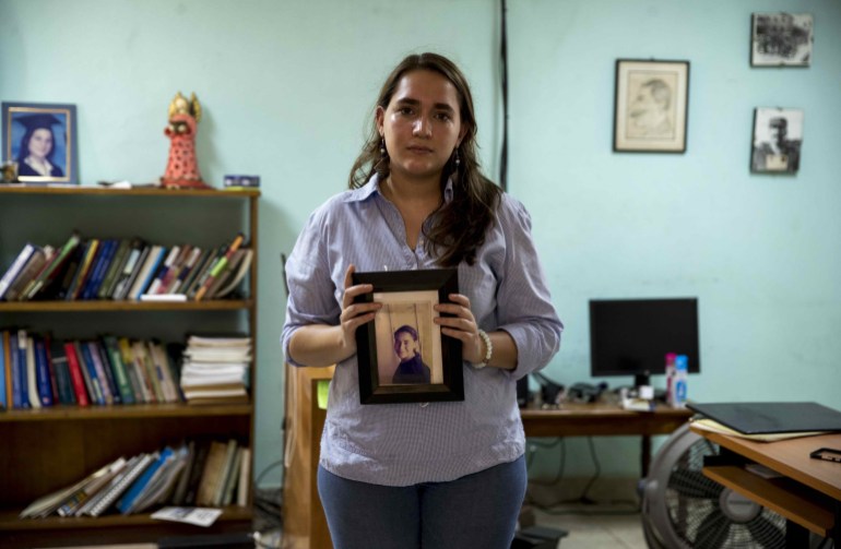 A woman holds a framed photo of Tamara Davila, her sister, in a room with bookshelves and a computer in the background.