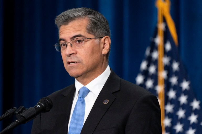 Seorang pria berjas gelap dan dasi biru muda, berkacamata, berdiri di depan bendera Amerika.