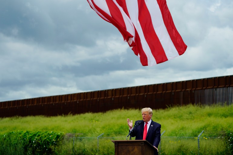 Donald Trump stands at a podium, speaking, as a US flag waves overhead and a grassy hill stands behind him, capped by a metal border barrier.