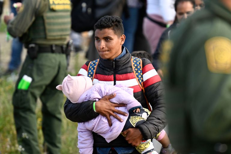 An asylum seeker carries his baby past US Border Patrol agents as they wait between the double fence along the US-Mexico border near Tijuana, Mexico, May 8, 2023