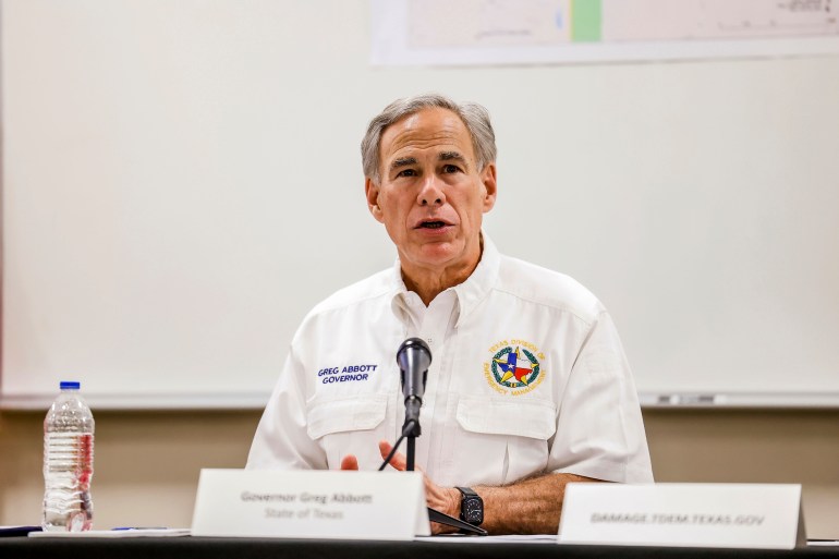 Governor Greg Abbott, dressed in a white uniform, sits at a panel table with a microphone in front of him.